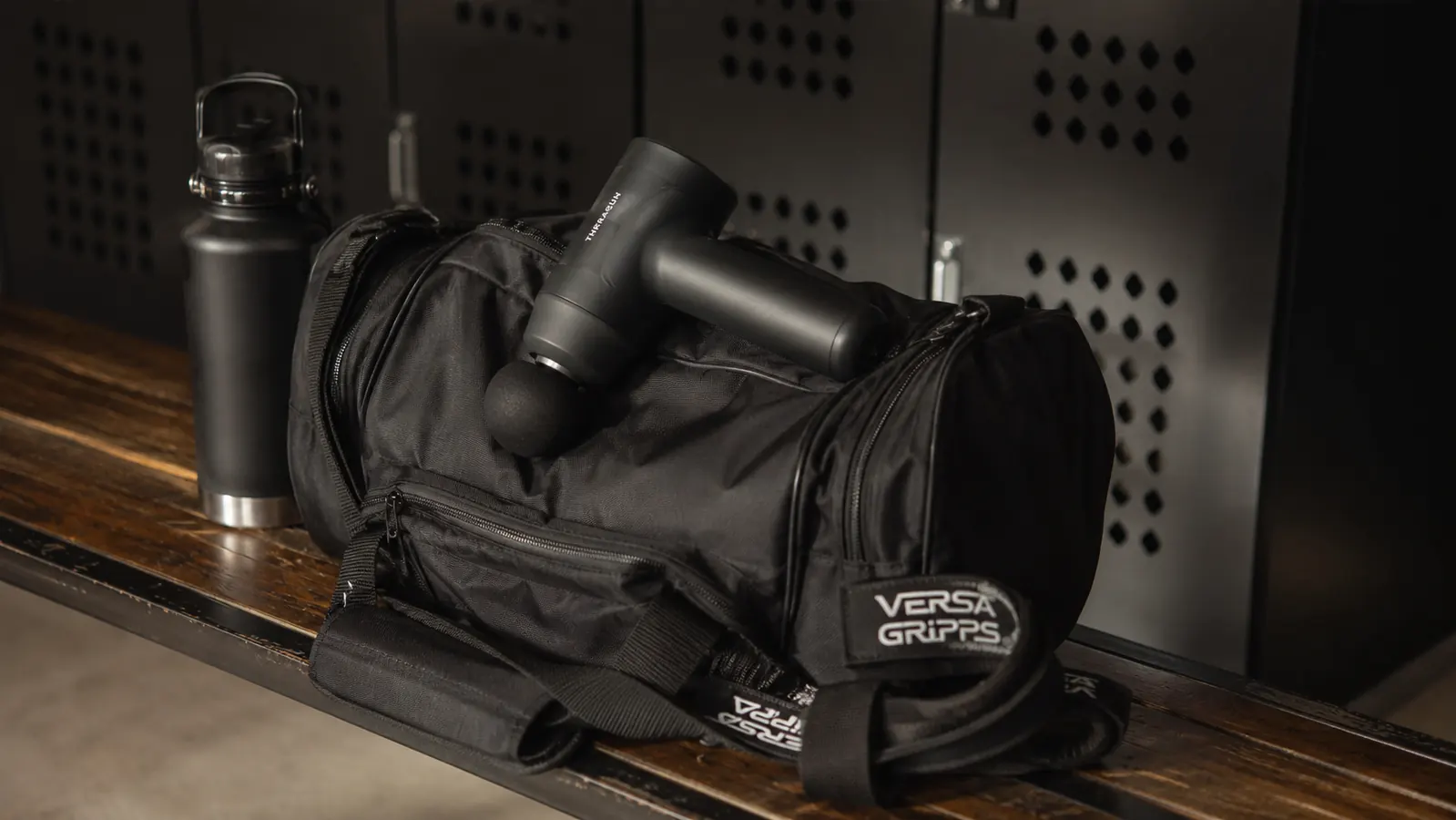 BOB AND BRAD Q2 Mini massage gun resting on a gym bag beside a water bottle and lifting straps on a wooden bench