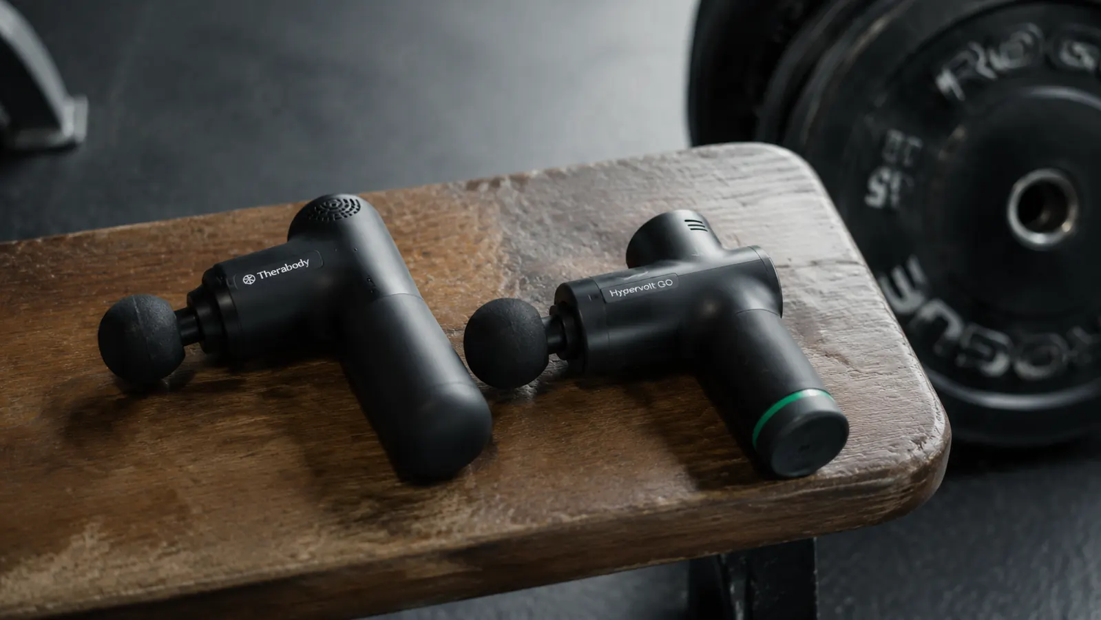 Two compact massage guns side by side on a gym bench with weights in the background