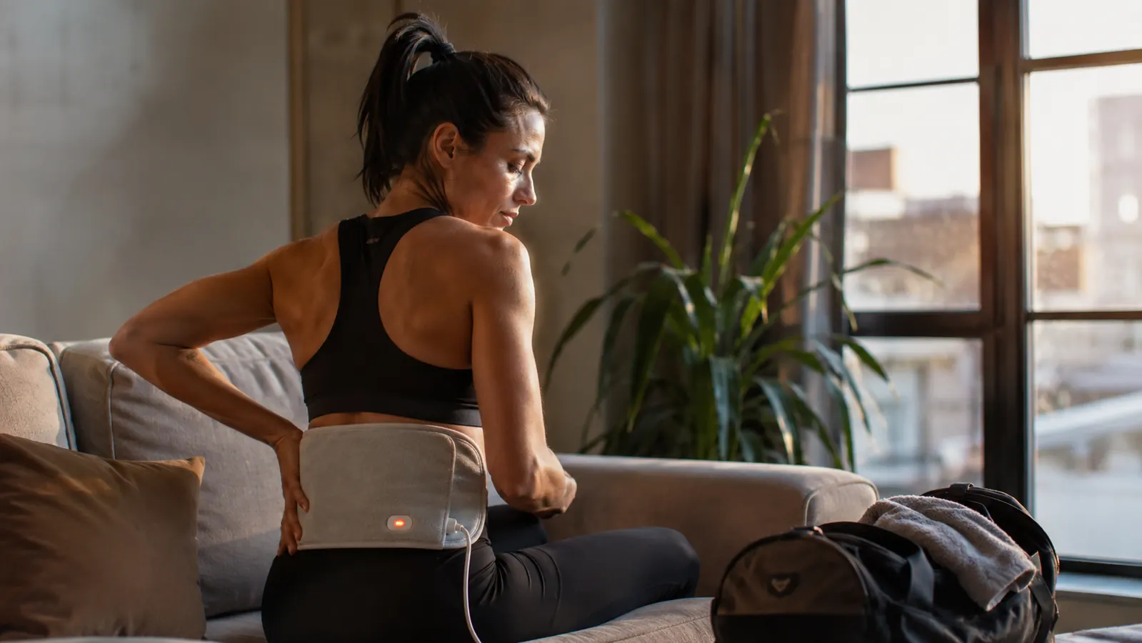 Person resting a heating pad on their lower back on a couch after a workout, gym bag visible in background